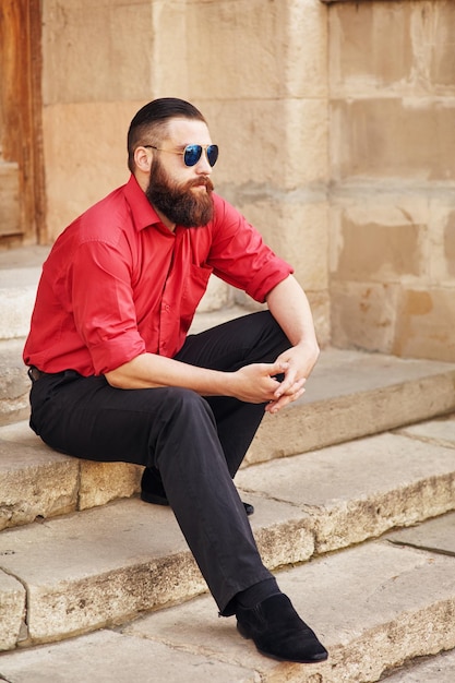 Giovane uomo che indossa occhiali da sole e vestito con camicia rossa e pantaloni neri seduto sotto il portico