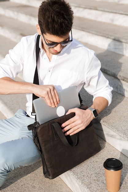 giovane uomo che indossa occhiali da sole, mettendo il computer portatile nella sua borsa, mentre è seduto sulle scale sulla strada della città