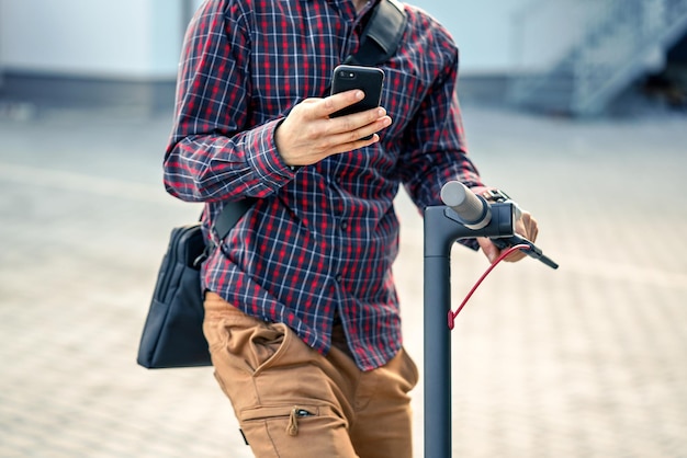 Giovane uomo che indossa una camicia appoggiata al manubrio dello scooter elettrico, tenendo in mano lo smartphone mobile, dettaglio del primo piano