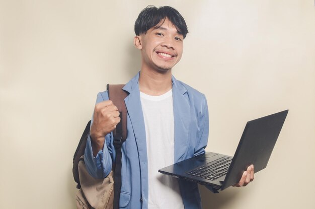 Giovane uomo che indossa una tuta da college con un gesto OK mentre trasporta il laptop su sfondo isolato