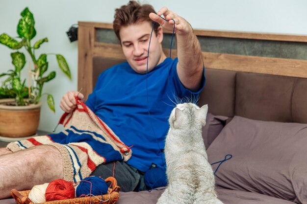 Giovane uomo che lavora a maglia un maglione a righe a casa Un simpatico gatto britannico gioca con un gomitolo di lana e interferisce con il lavoro a maglia del suo proprietario