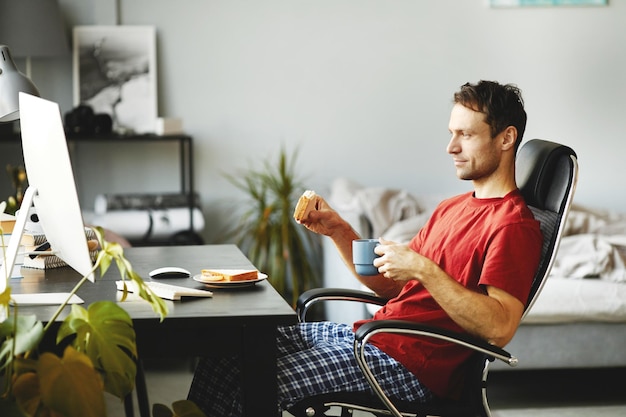 Giovane uomo che mangia panino e beve caffè mentre è seduto su una sedia davanti al computer e watchi