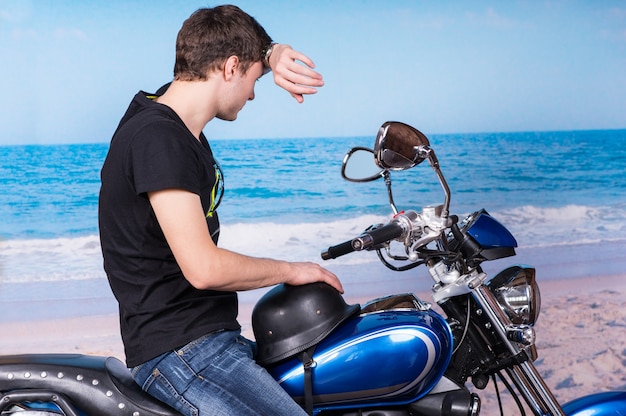 Giovane uomo che pulisce la fronte con il polso seduto su una motocicletta blu classica in spiaggia e guardando in lontananza