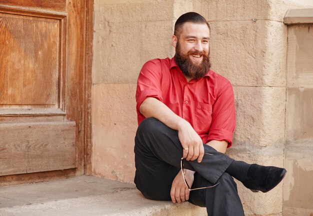 Giovane uomo che ride vestito con camicia rossa e pantaloni neri seduto sotto il portico