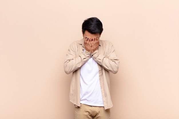 Giovane uomo cinese che si sente triste, frustrato, nervoso e depresso, coprendosi il viso con entrambe le mani, piangendo contro la parete di colore piatto