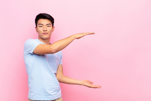 Giovane uomo cinese che tiene un oggetto con entrambe le mani sullo spazio laterale della copia, mostrando, offrendo o pubblicizzando un oggetto sulla parete di colore piatto