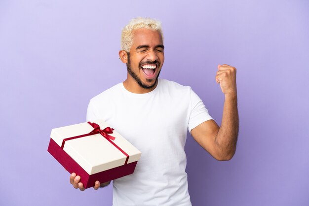 Giovane uomo colombiano che tiene un regalo isolato su sfondo viola che celebra una vittoria