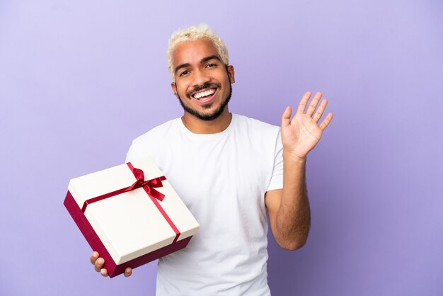 Giovane uomo colombiano che tiene un regalo isolato su sfondo viola salutando con la mano con espressione felice