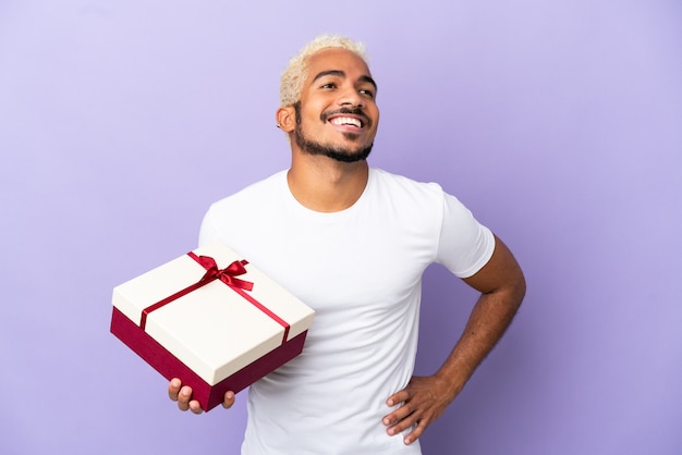 Giovane uomo colombiano in possesso di un regalo isolato su sfondo viola in posa con le braccia all'anca e sorridente