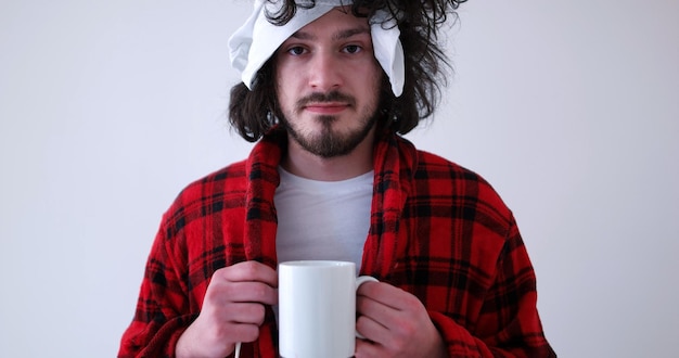Giovane uomo con influenza e febbre avvolta in possesso di una tazza di tè curativo isolato su bianco