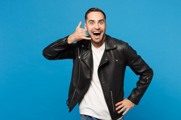 Giovane uomo con la barba lunga in giacca nera t-shirt bianca che guarda la macchina fotografica facendo il gesto del telefono come dice chiamami indietro isolato sul ritratto dello studio del fondo della parete blu. Concetto di stile di vita delle persone Mock up copy space