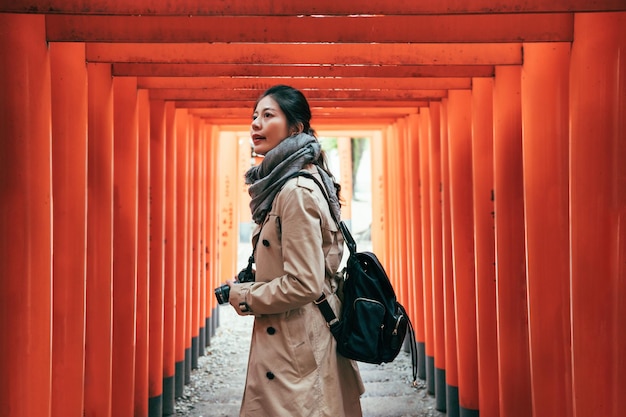 giovane uomo con lente turistica che tiene la macchina fotografica che cammina attraverso i cancelli rossi di torii tornando indietro guardando il cielo. Il fotografo zaino in spalla ama l'hobby che scatta foto fantastiche durante il viaggio.