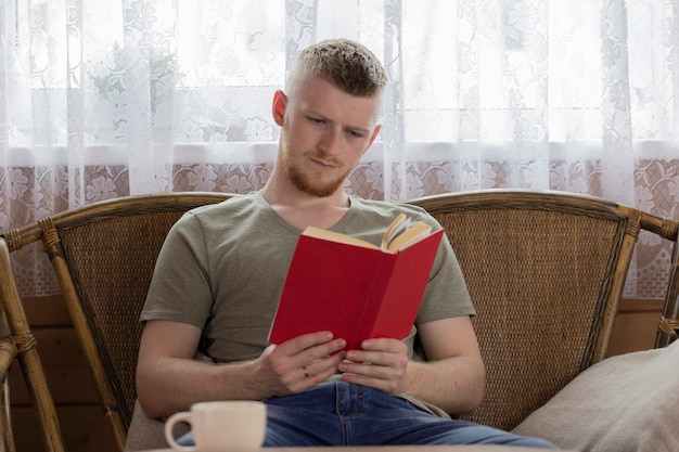 Giovane uomo concentrato a leggere il libro con copertina rossa sul banco di vimini in casa di campagna in legno