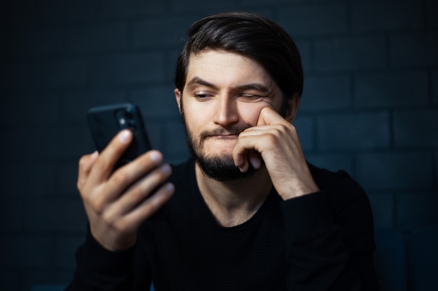 Giovane uomo confuso che guarda nello smartphone sullo sfondo del muro di mattoni nero.