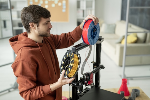 Giovane uomo creativo cambiando bobina con filamento mentre è in piedi dalla stampante 3d prima di stampare oggetti di vari colori