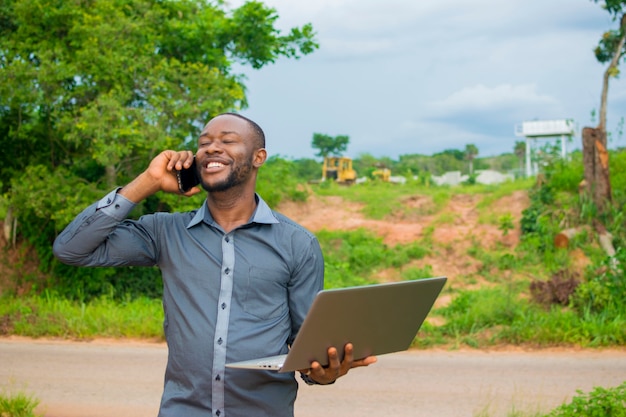 Giovane uomo d'affari africano bello in piedi su una terraferma, sentendosi sovreccitato mentre tiene il suo laptop e fa chiamate con il suo cellulare.