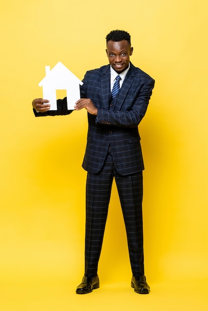 Giovane uomo d'affari africano come agente immobiliare che tiene il modello di ritaglio della casa in sfondo giallo isolato per studio