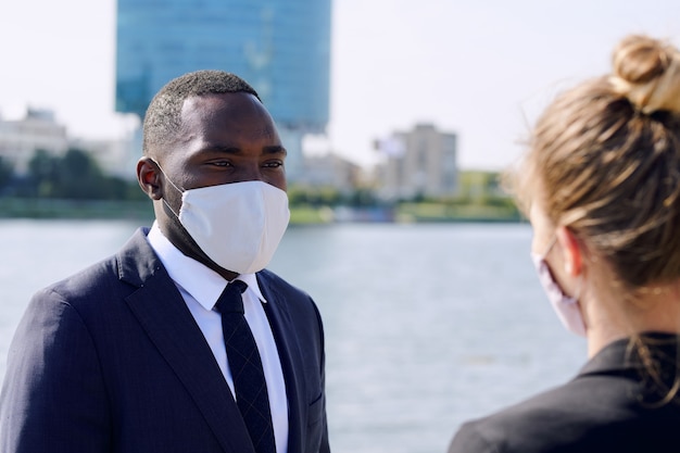 Giovane uomo d'affari africano in maschera protettiva e abbigliamento formale che guarda la sua collega durante la discussione sui punti di lavoro all'aperto