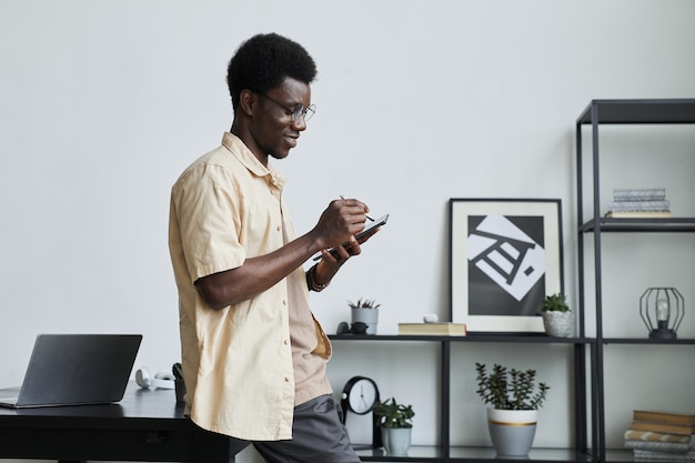 Giovane uomo d'affari africano in piedi vicino al suo posto di lavoro in ufficio e sorridente che comunica online ...