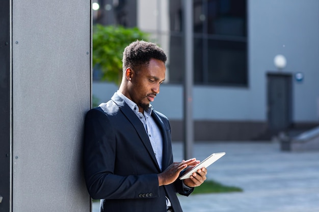 Giovane uomo d'affari afroamericano in tailleur formale in piedi lavorando con tablet in mani su sfondo moderno edificio per uffici al di fuori. Uomo che utilizza smartphone o telefono cellulare all'aperto strada della città