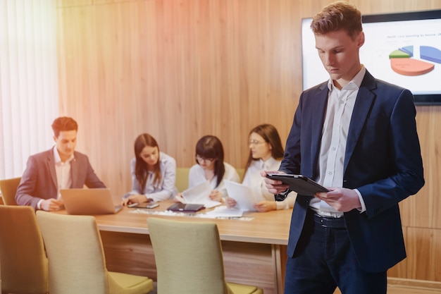 Giovane uomo d'affari alla moda che indossa una giacca e una camicia sullo sfondo di un ufficio di lavoro con persone che lavorano con un tablet