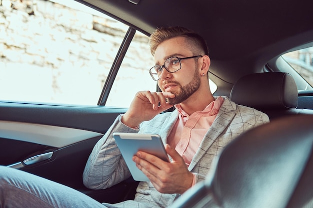 Giovane uomo d'affari alla moda di successo in abito grigio e camicia rosa, pensieroso con un tablet in mano, in sella a un sedile posteriore in un'auto di lusso.