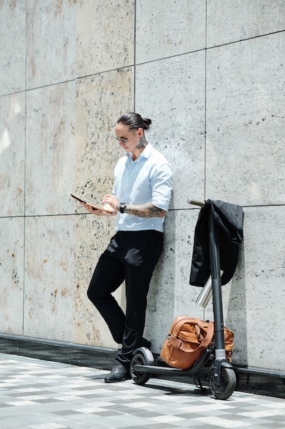 Giovane uomo d'affari alla moda in piedi all'aperto accanto al suo scooter e leggere il documento sulla tavoletta digitale