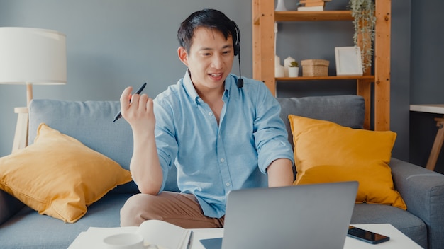 Giovane uomo d'affari asiatico che indossa le cuffie utilizzando laptop e parla con i colleghi sul piano in videochiamata