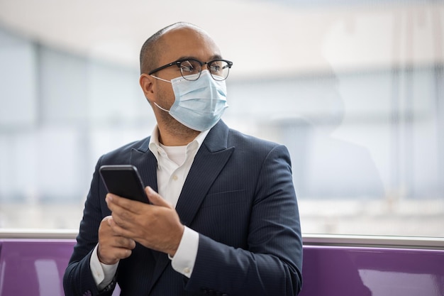 Giovane uomo d'affari asiatico che indossa una maschera di protezione utilizzando lo smartphone seduto in skytrain. Uomo d'affari nel viaggio urbano della città in treno sopraelevato.