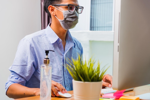 Giovane uomo d'affari asiatico che indossa una maschera protettiva con gel disinfettante accanto a lui lavora da casa in remoto online con PC computer mette in quarantena la malattia coronavirus o COVID-19