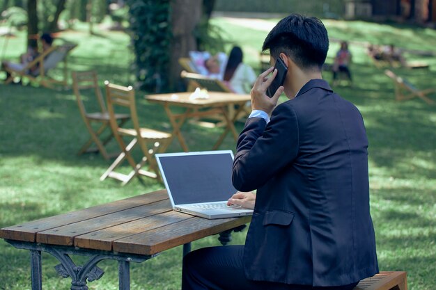 Giovane uomo d'affari asiatico con laptop e notebook che lavorano nel parco con chat video sul computer portatile con un collega mentre si è seduti all'aperto in giardino. Affari e tecnologia