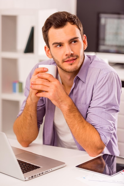 Giovane uomo d'affari attraente in abiti casual che beve caffè e sogna mentre lavora in ufficio