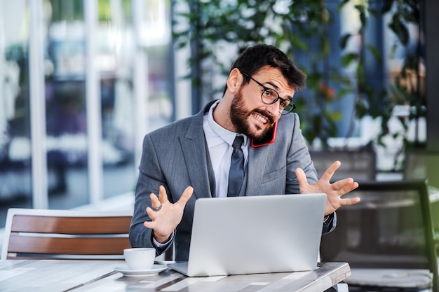 Giovane uomo d'affari barbuto caucasico attraente che si siede sulla terrazza del caffè, tenendo lo smart phone sulla spalla e convincendo il cliente ad avere una collaborazione con lui.