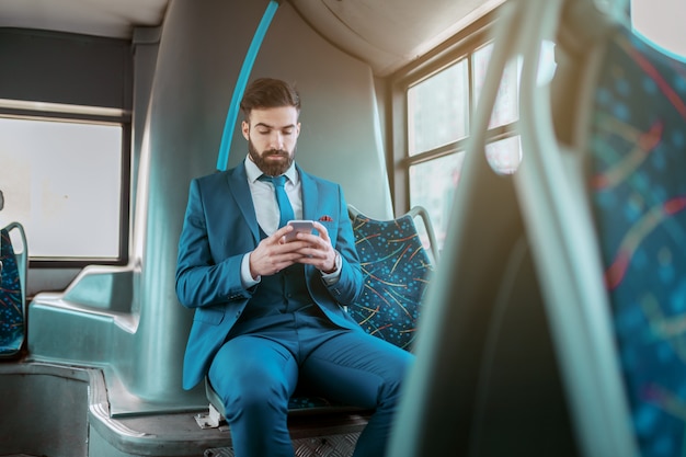 Giovane uomo d'affari barbuto caucasico serio attraente in vestito blu che si siede in autobus pubblico e utilizzando smart phone.