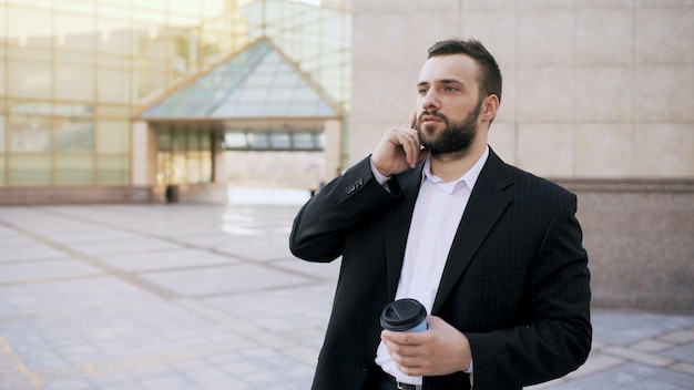 Giovane uomo d'affari barbuto che parla allo smartphone con una tazza di caffè vicino agli edifici per uffici all'aperto