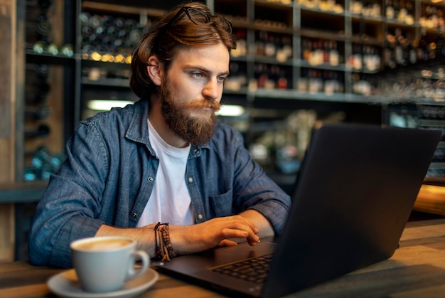 Giovane uomo d'affari barbuto concentrato che lavora da un computer portatile in un caffè