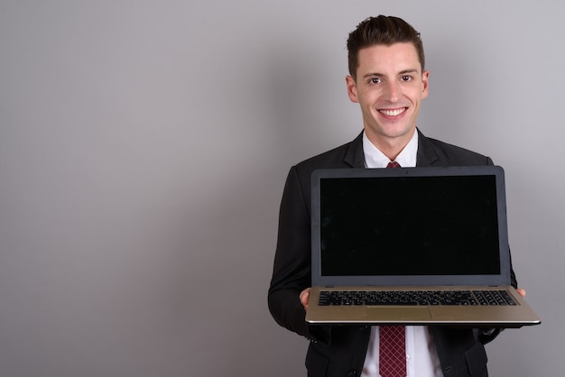 giovane uomo d'affari bello che indossa tuta mentre si tiene il computer portatile su grigio