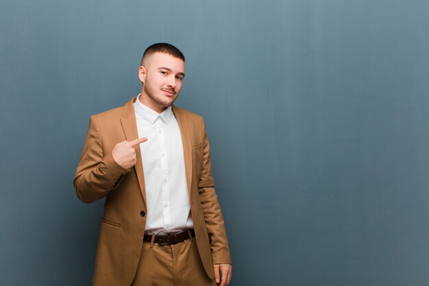 Giovane uomo d'affari bello che sembra fiero, sicuro e felice, sorridendo e indicando se stesso o facendo il segno di numero uno contro la parete piana