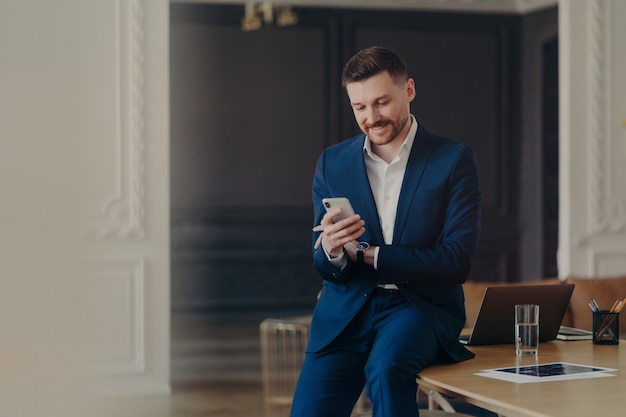 Giovane uomo d'affari bello in abito blu scuro formale che guarda lo smartphone e sorride mentre legge i messaggi, seduto casualmente sulla scrivania in un elegante ufficio minimalista o lavora da casa