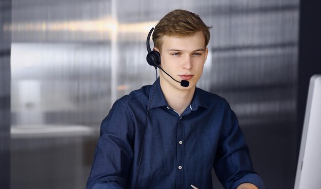 Giovane uomo d'affari biondo che utilizza auricolare e computer al lavoro. Avviare un'attività significa lavorare sodo e fuori tempo per raggiungere il successo.