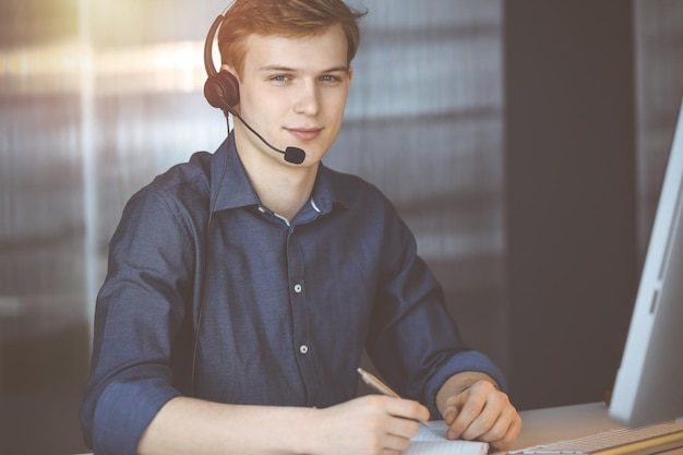 Giovane uomo d'affari biondo che utilizza auricolare e computer in un ufficio oscurato, bagliore di luce sullo sfondo. Avviare un'impresa significa lavorare sodo.