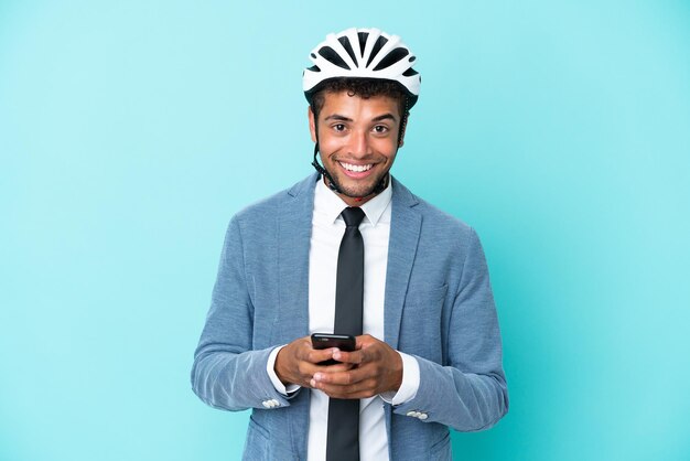 Giovane uomo d'affari brasiliano con casco da bici isolato su sfondo blu che invia un messaggio con il cellulare