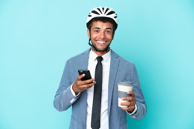 Giovane uomo d'affari brasiliano con casco da bici isolato su sfondo blu che tiene caffè da portare via e un cellulare