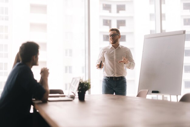 Giovane uomo d'affari casual che presenta un nuovo progetto ai partner sulla lavagna in un ufficio moderno che si riunisce contro la grande finestra panoramica. Concetto di vita in ufficio.