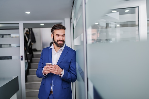 Giovane uomo d'affari caucasico bello in vestito che sta nel corridoio della ditta di broker e che per mezzo dello Smart Phone.