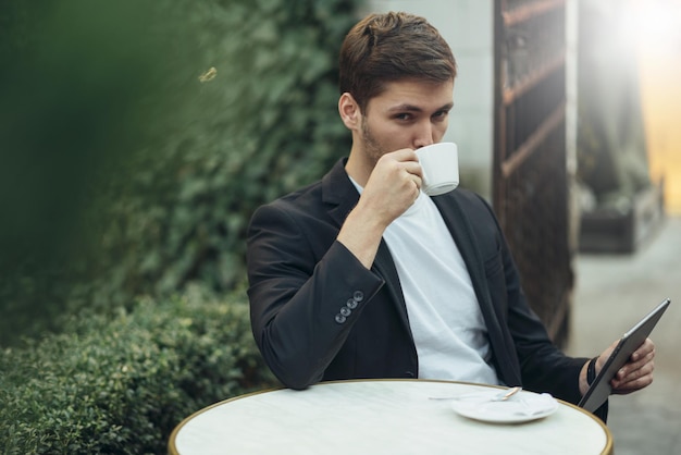 Giovane uomo d'affari caucasico che controlla il feed sul suo dispositivo e beve caffè caldo