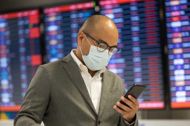 Giovane uomo d'affari che indossa una maschera per proteggere l'influenza coronavirus Covid-19. Trascina i bagagli camminando nel terminal. Turista nel terminal dell'aeroporto tramite smartphone effettua il check-in all'orario di volo informativo.