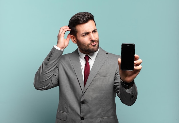 Giovane uomo d'affari che si sente perplesso e confuso grattandosi la testa e guardando di lato e mostrando il suo schermo del telefono
