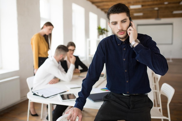 Giovane uomo d'affari con la barba in camicia scura che guarda premurosamente da parte mentre parla al cellulare in ufficio con i colleghi sullo sfondo