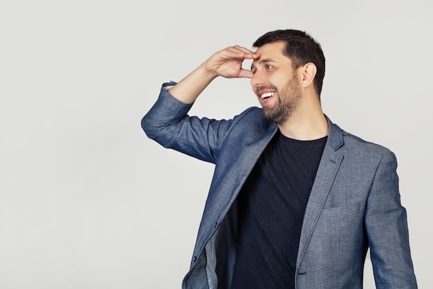 Giovane uomo d'affari con la barba in una giacca, molto felice e sorridente, guardando lontano con la mano sopra la sua testa.